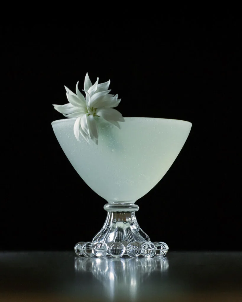 a white cocktail in a coupe glass with a white flower garnish.