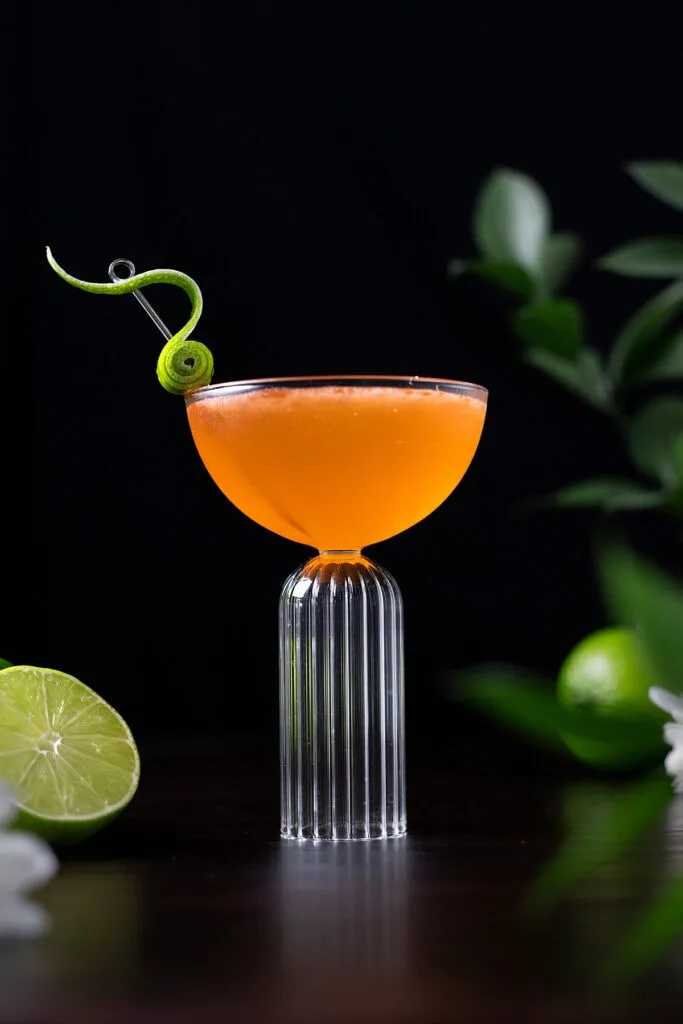 orange cocktail in a coupe glass with a fancy lime peel garnish.