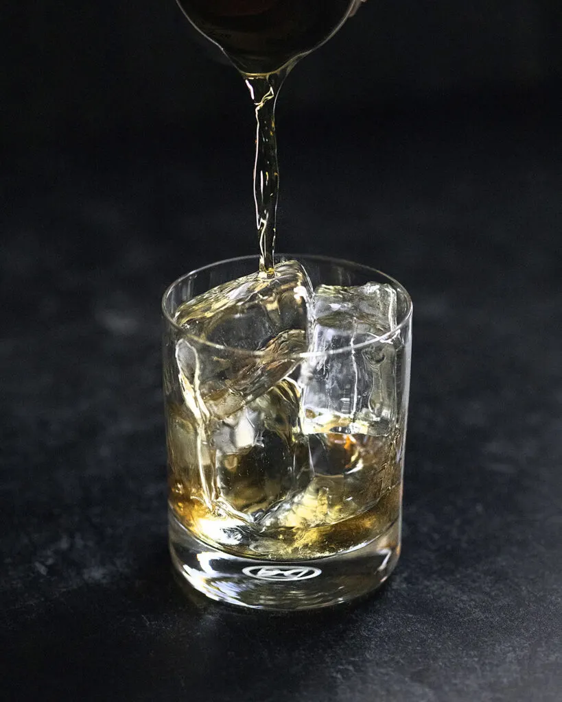 pouring whiskey over clear ice cubes in a rocks glass.
